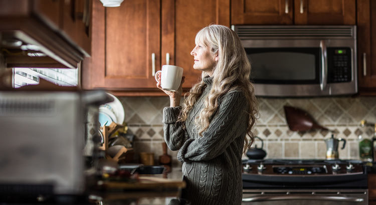 Photo of an older woman standing in kitchen drinking coffee