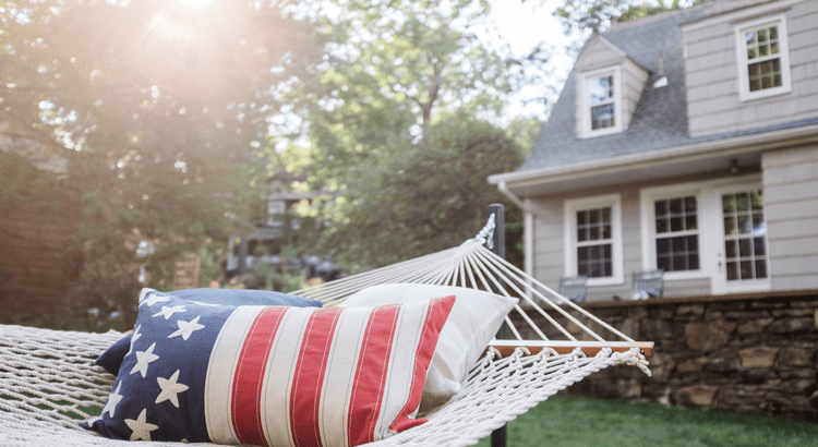A hammock with an American flag pillow in a sunny backyard, with a cozy house and green trees in the background.