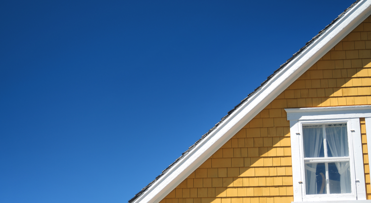 A close-up view of the upper portion of a yellow house with a gable roof and white trim against a clear blue sky. A single window with white curtains is visible.