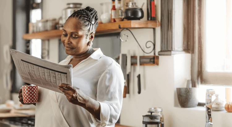 A woman in a white blouse reads a newspaper while holding a red patterned mug in a cozy, sunlit kitchen.