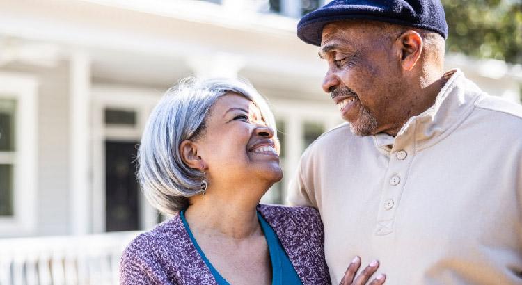 A joyful elderly couple smiles warmly at each other, standing outside their home, embracing lovingly in the sunlight.