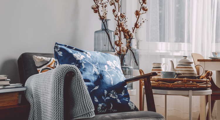 Cozy living room corner with a grey chair, blue floral pillow, knit blanket, and a tea set on a small table.